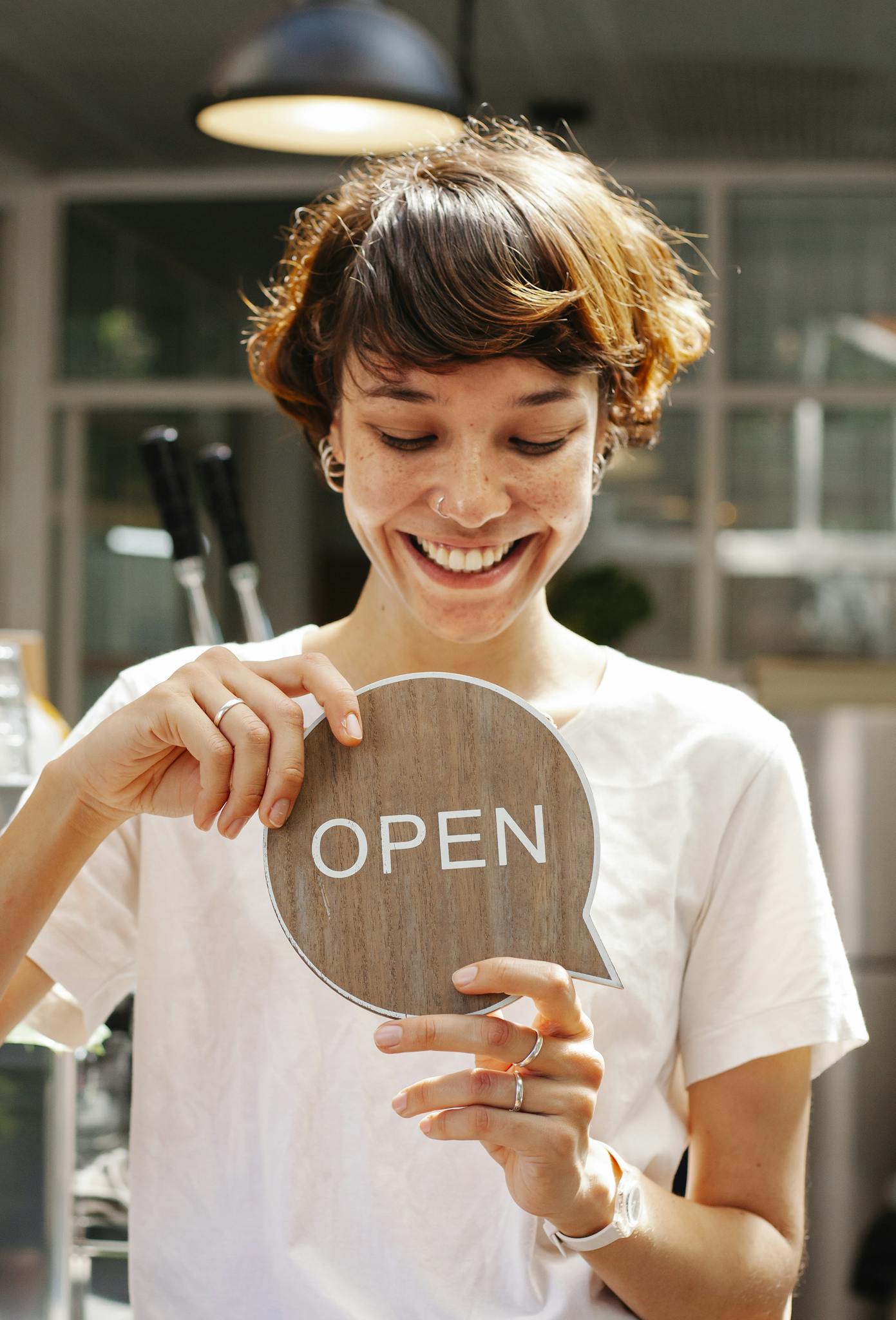 Through glass of happy female worker holding wooden Open sign working in modern cafe with glowing lamp inside