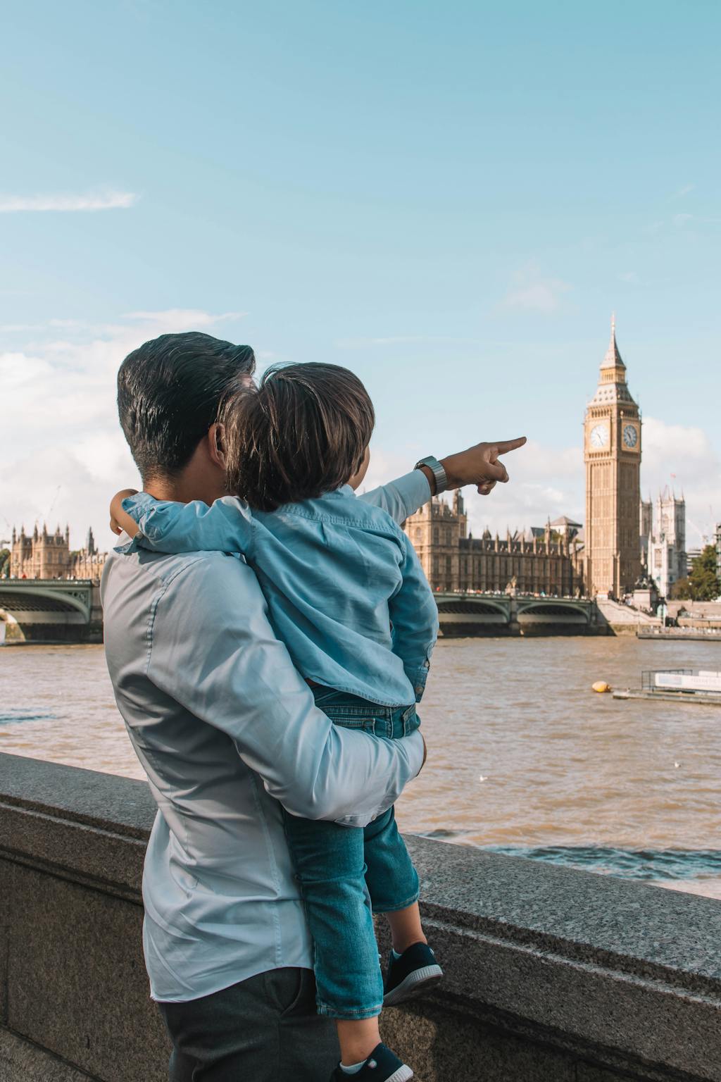 Father holds child as they point towards Big Ben on a sunny day in London.